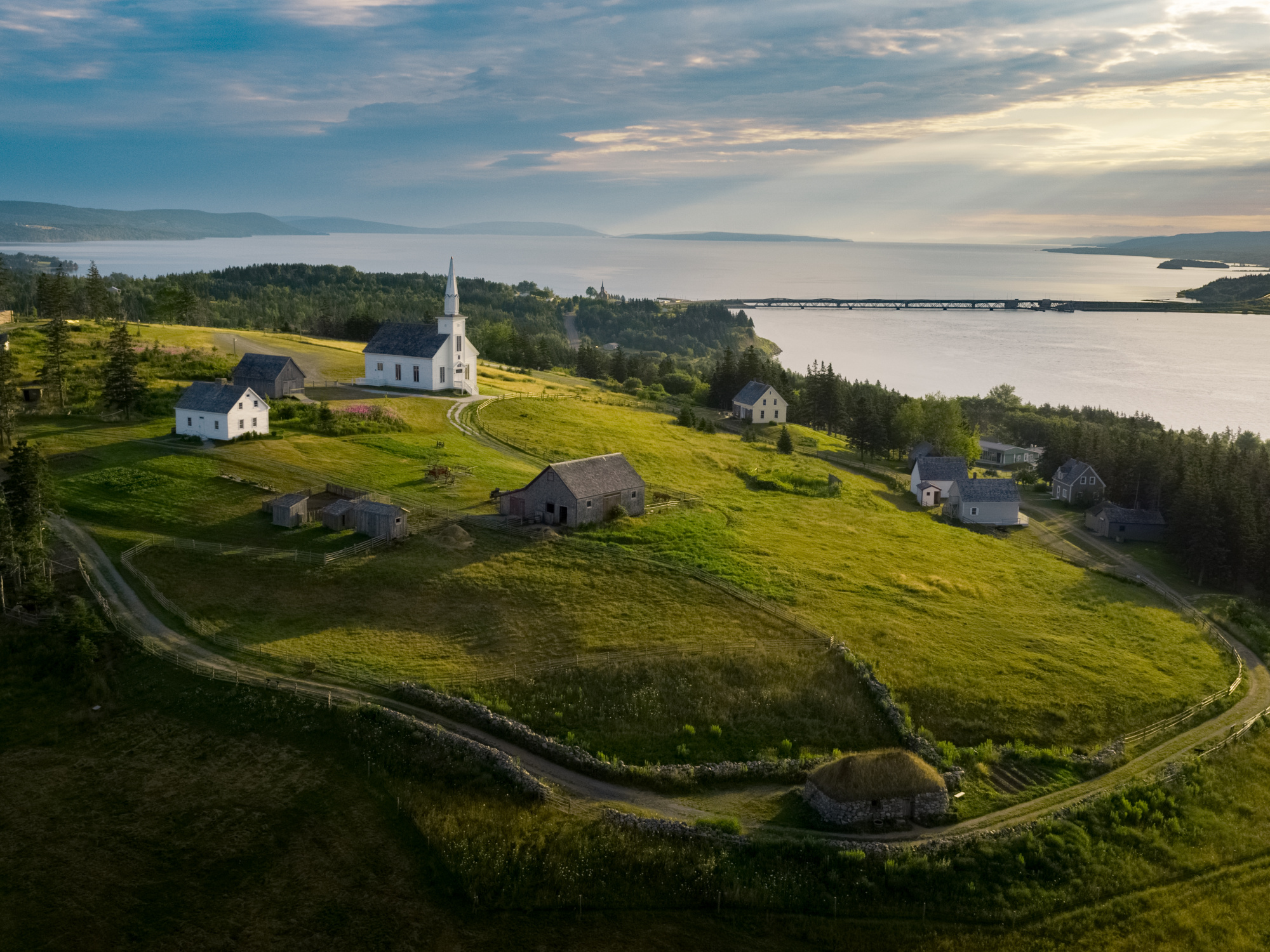 An aerial view captures the Highland Village Museum in Iona, Nova Scotia, showcasing a collection of historic buildings scattered across a green hillside overlooking a body of water under a partly cloudy sky. A white church with a steeple stands prominently among the other smaller, older structures.