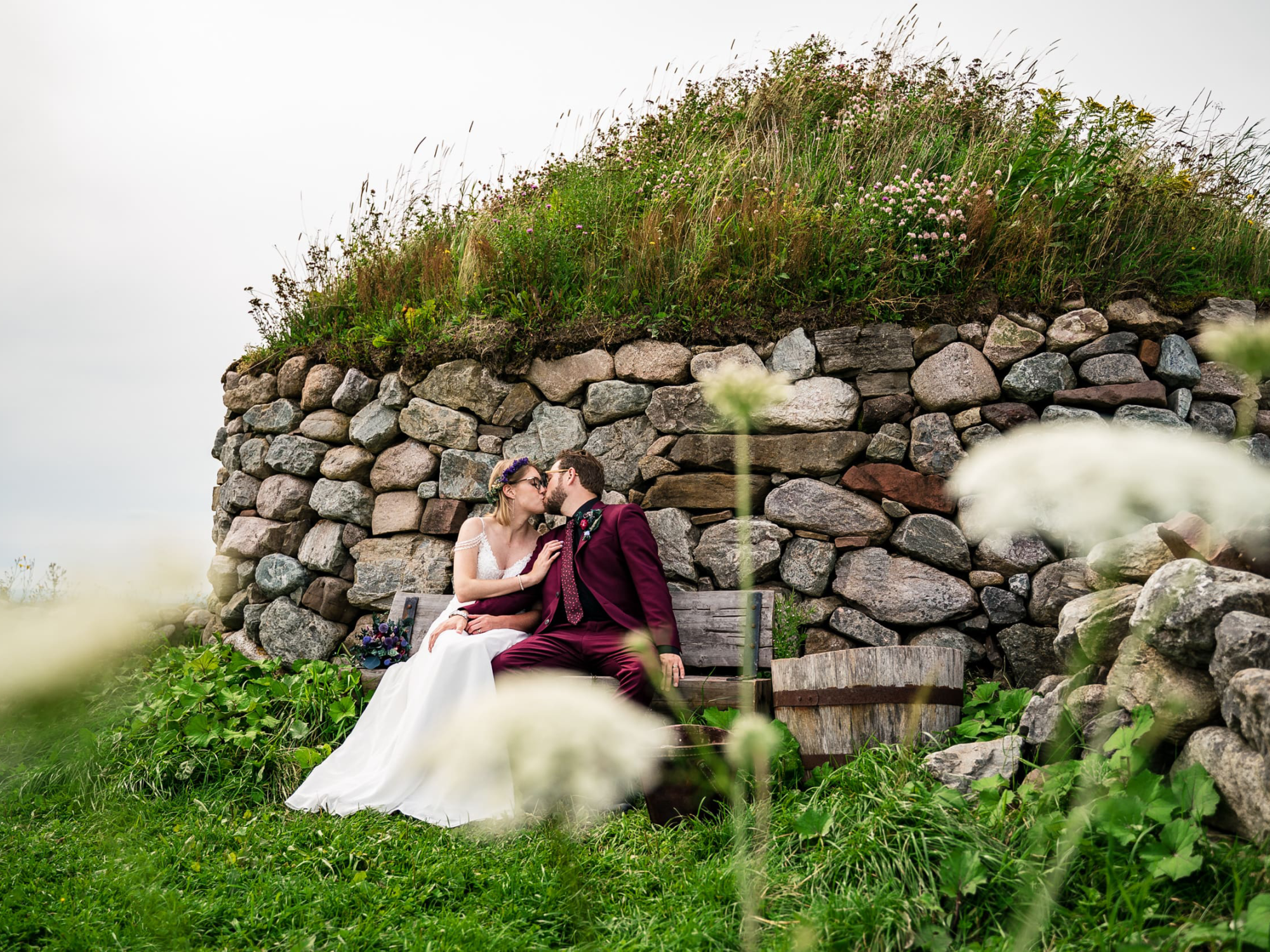 A wedding couple, with the bride in a white gown and groom in a maroon suit, sharing a kiss on a wooden bench. They are seated in front of a historic Scottish blackhouse dwelling at the Highland Village Museum, which features rugged stone walls and a thick roof of grass and wildflowers. Blurred white wildflowers frame the scene in the foreground.