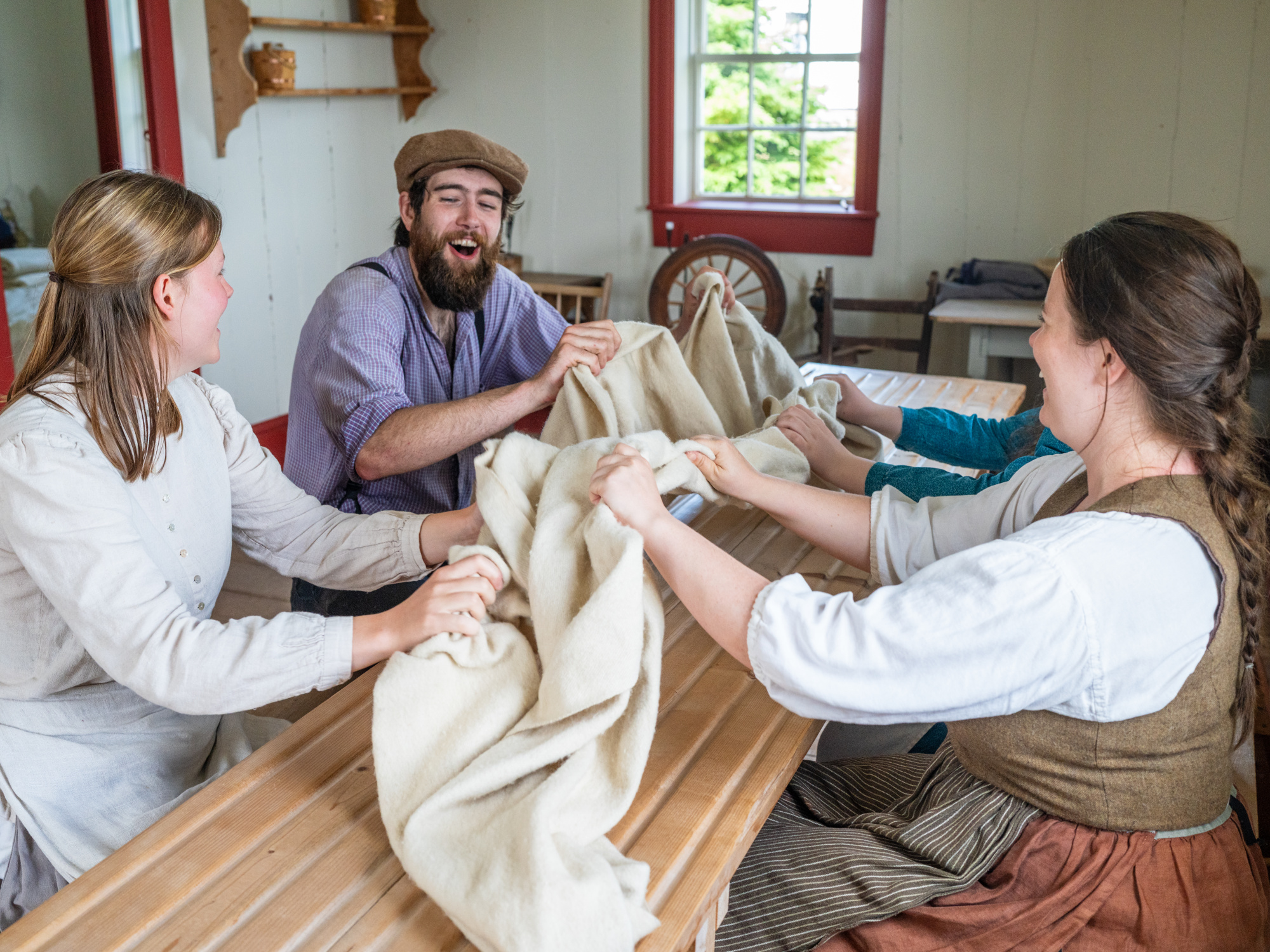 Four costumed interpreters sit around a long wooden table inside a historic room, laughing while participating in a traditional milling frolic. They are gathered closely, using their hands to rhythmically pull and bunch a large piece of cream-colored woven wool cloth across the surface of the slatted table.
