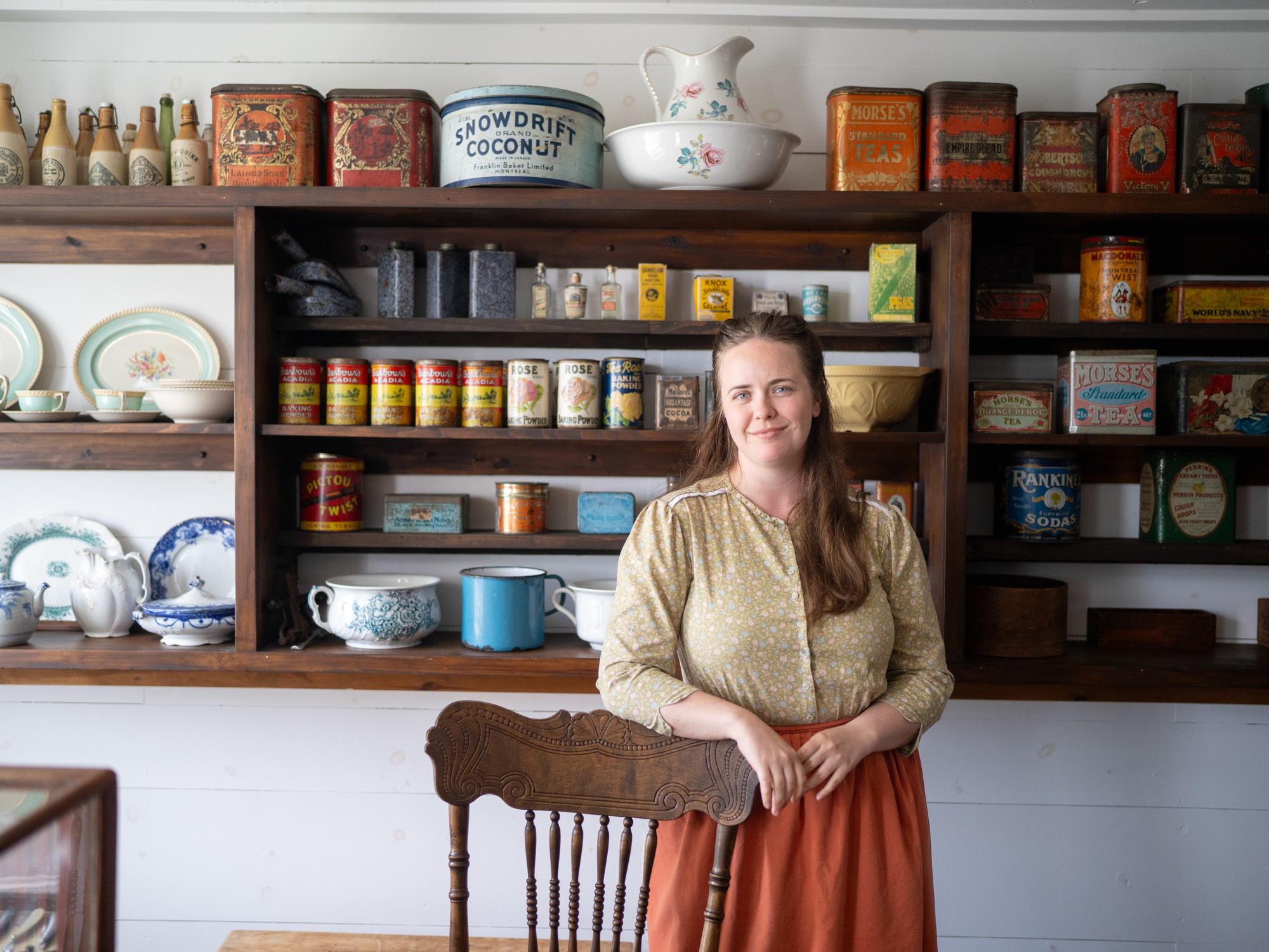 A costumed interpreter stands smiling behind a historic wooden chair inside a vintage general store setting. She wears a long-sleeved, yellow patterned blouse and a rust-orange skirt. Behind her, floor-to-ceiling wooden shelving units are filled with a dense display of antique packaging, including colorful tin containers for tea, coffee, and coconut, rows of small glass spice bottles, canned goods, and a collection of ceramic plates, teapots, and wash basins.