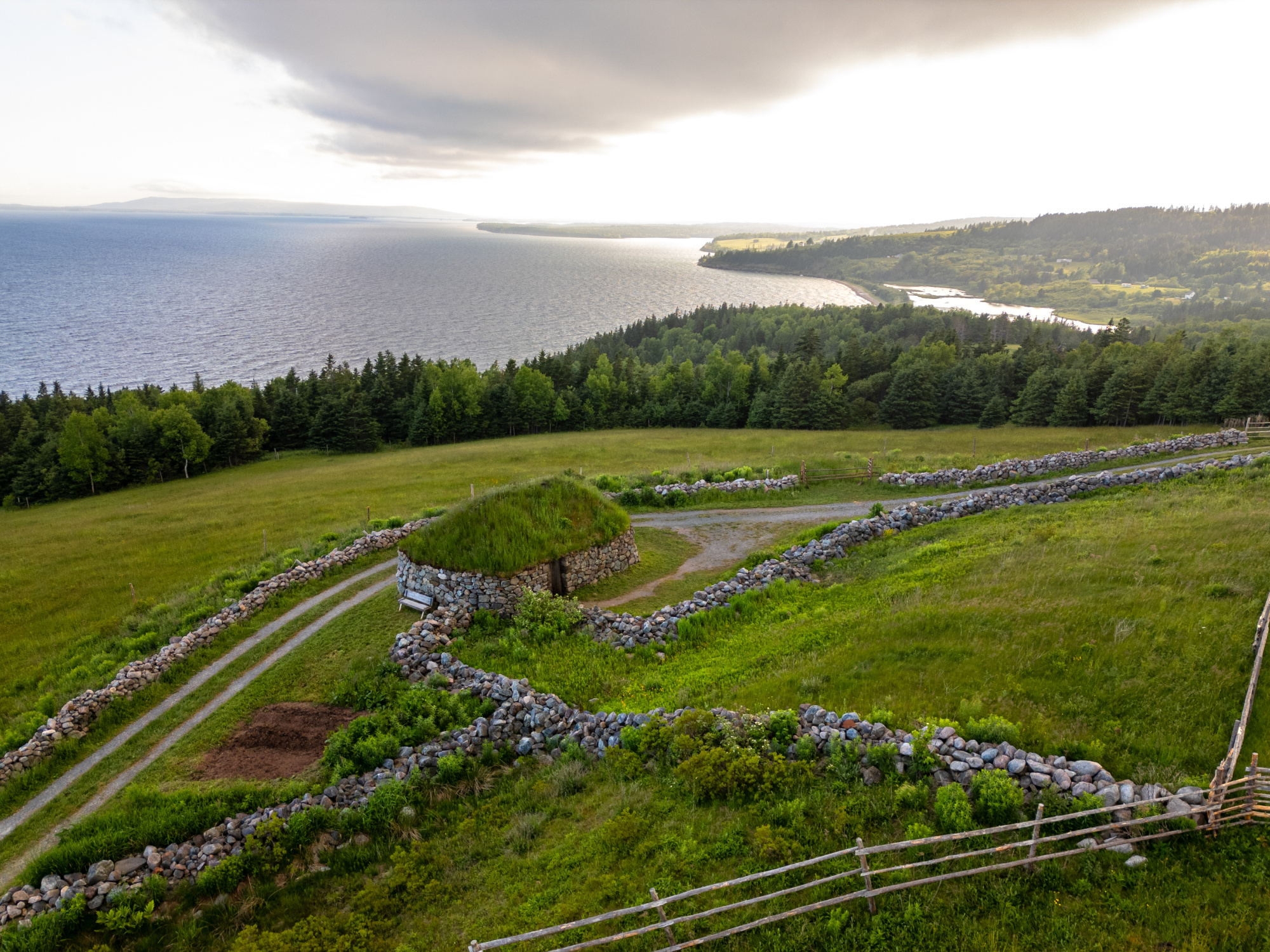 An elevated, wide-angle view looking down at a traditional sod-roofed stone building on a grassy hillside at Highland Village Museum. The circular stone structures are nestled within a network of low, rustic stone walls and wooden fences that section off the bright green fields. A double-track dirt path runs alongside the property, leading down the hillside toward a thick treeline of evergreens.