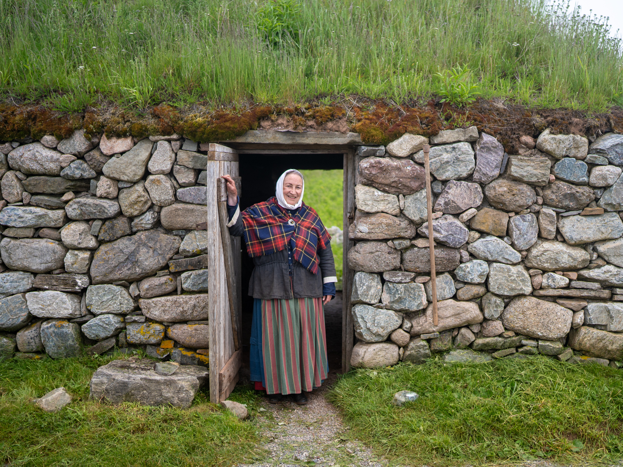 A smiling costumed interpreter stands in the open wooden doorway of a traditional sod-roofed stone building at Highland Village Museum. The building features low walls constructed from uneven fieldstones topped with green grass and moss. The interpreter is wearing a white headscarf, a dark jacket, a red, blue, and green tartan shawl pinned at the chest, and a long striped skirt. A small dirt path leads up to the entrance.
