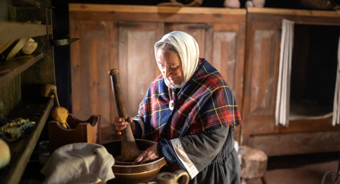 A close-up interior view of a costumed interpreter inside a traditional building at Highland Village Museum. The woman wears a white headscarf and a red, blue, and yellow tartan shawl pinned at her chest while using a large wooden pestle to grind ingredients in a bowl on a rustic counter. The dimly lit historic room features large wooden storage cupboards, a built-in bed box with a white curtain, pottery jars, and various period kitchen utensils.