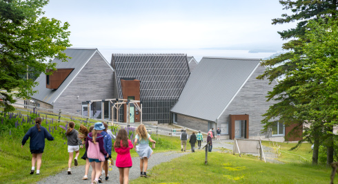 A view from a grassy hillside path looking down toward the modern visitor centre at Highland Village Museum. A group of children is walking down the gravel pathway in the foreground, with other visitors visible further ahead. The building features a contemporary design with multi-peaked metal roofs, grey wood siding, and rusted metal accents. A hillside field with purple lupins stands to the left, and a vast body of water is visible under an overcast sky in the background.