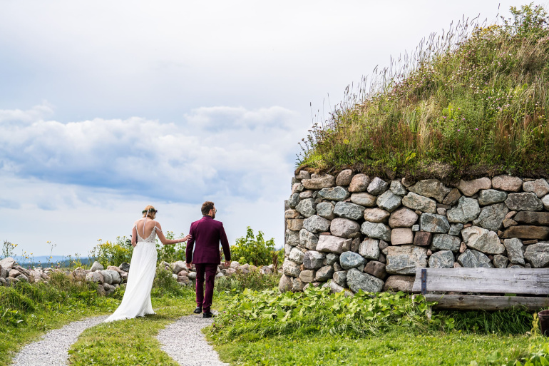 A wedding couple walks down a gravel path at the Highland Village Museum toward the Bras d'Or Lake, passing a traditional Scottish blackhouse with a sod roof.
