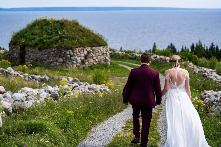 A wedding couple walks down a gravel path at the Highland Village Museum toward the Bras d'Or Lake, passing a traditional Scottish blackhouse with a sod roof