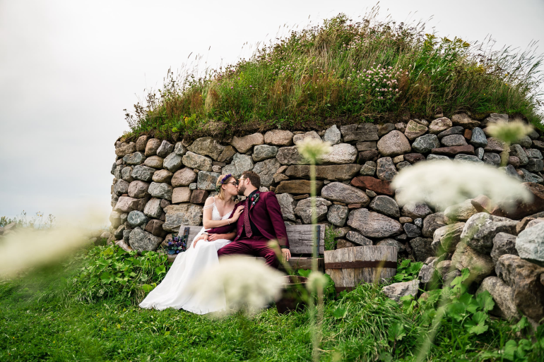 A wedding couple, with the bride in a white gown and groom in a maroon suit, sharing a kiss on a wooden bench. They are seated in front of a historic Scottish blackhouse dwelling at the Highland Village Museum, which features rugged stone walls and a thick roof of grass and wildflowers. Blurred white wildflowers frame the scene in the foreground.