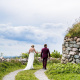 A wedding couple walks down a gravel path at the Highland Village Museum toward the Bras d'Or Lake, passing a traditional Scottish blackhouse with a sod roof.