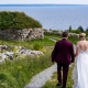 A wedding couple walks down a gravel path at the Highland Village Museum toward the Bras d'Or Lake, passing a traditional Scottish blackhouse with a sod roof