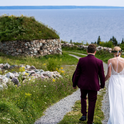 A wedding couple walks down a gravel path at the Highland Village Museum toward the Bras d'Or Lake, passing a traditional Scottish blackhouse with a sod roof