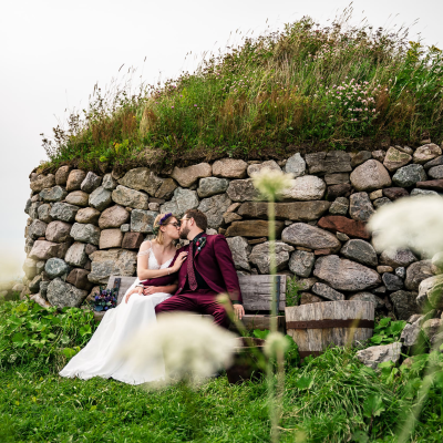 A wedding couple, with the bride in a white gown and groom in a maroon suit, sharing a kiss on a wooden bench. They are seated in front of a historic Scottish blackhouse dwelling at the Highland Village Museum, which features rugged stone walls and a thick roof of grass and wildflowers. Blurred white wildflowers frame the scene in the foreground.