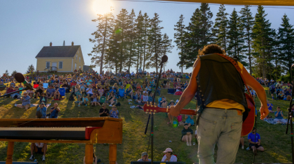 A musician seen from behind plays a guitar on an outdoor stage, facing a large crowd seated on a grassy hill. In the background, a historic yellow house sits near a line of tall evergreen trees under a bright, sunny sky.