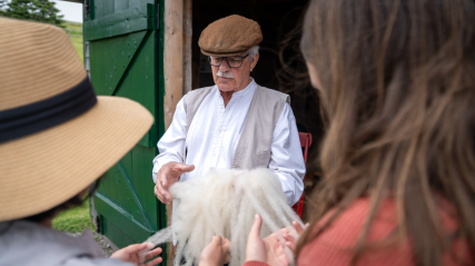 A historical interpreter shows raw wool to visitors outside a wooden building with green doors.