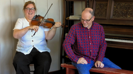 A smiling woman in a polka-dot blouse plays the fiddle while a man in a red and blue plaid shirt sits on a bench next to an upright piano, listening or preparing to accompany her.