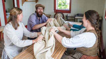 Four costumed interpreters sit around a long wooden table inside a historic room, laughing while participating in a traditional milling frolic (luadh). They are gathered closely, using their hands to rhythmically pull and bunch a large piece of cream-colored woven wool cloth across the surface of the slatted table. In the background, the room features white walls with red window trim, a spinning wheel, and wooden shelving.
