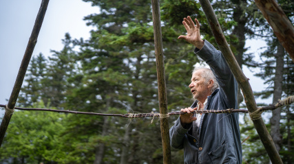Side profile of an older man with grey hair wearing a grey jacket, gesturing with an open hand raised as he stands behind a horizontal wooden branch lashed to vertical poles with twine. Evergreen forest fills the background under an overcast sky.