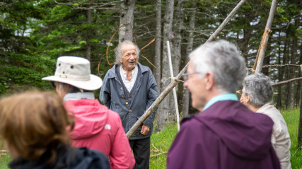 An outdoor, medium shot captures an older man with grey hair and a weathered face speaking warmly to a small group of visitors, who are viewed from behind and slightly out of focus in the foreground.  The speaker stands on a grassy patch at the edge of a dense forest, smiling as he talks.
