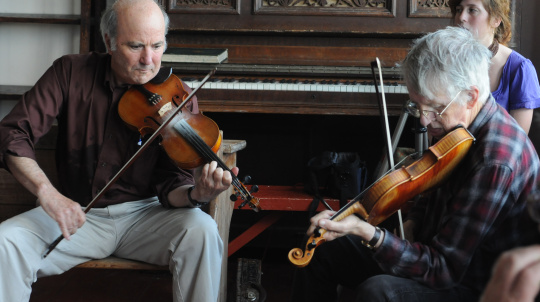 Musicians playing fiddles in front of a piano.