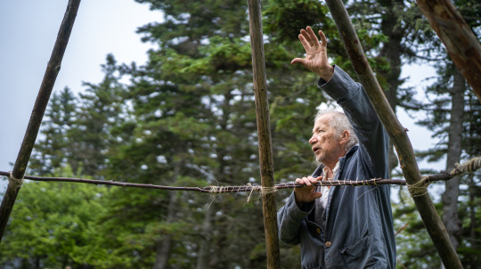 Side profile of an older man with grey hair wearing a grey jacket, gesturing with an open hand raised as he stands behind a horizontal wooden branch lashed to vertical poles with twine. Evergreen forest fills the background under an overcast sky.